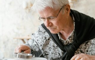 Ein älterer Mann mit Brille nutzt eine Lupe zum Lesen, da seine Sehschärfe nachgelassen hat – ein mögliches Anzeichen für Grauen Star. Verschwommenes Sehen und Schwierigkeiten beim Lesen gehören zu den frühen Symptomen dieser Augenerkrankung
